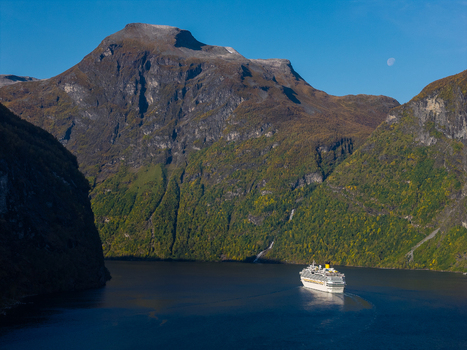 Aerial view of a cruise ship sailing through Geirangerfjord surrounded by steep mountains with autumn foliage under a clear blue sky in Geiranger More og Romsdal Norway. Aerial view of a cruise ship sailing through Geirangerfjord surrounded by steep mountains with autumn foliage under a clear blue sky in Geiranger More og Romsdal Norway.