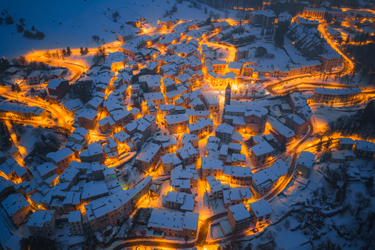 Aerial view of the snow covered town at night with warm street lights illuminating winding roads and historic buildings in Rivisondoli Abruzzo Italy.
