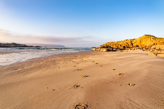 View of the sandy beach at Cloughglass with golden rocky outcrops gentle waves and footprints in the sand under a clear blue sky in Cloughglass County Donegal Ireland. View of the sandy beach at Cloughglass with golden rocky outcrops gentle waves and footprints in the sand under a clear blue sky in Cloughglass County Donegal Ireland.