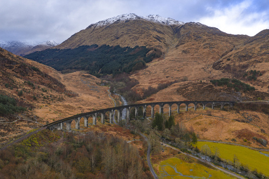 Aerial view of the historic Glenfinnan Viaduct with its iconic curved arches spanning a rugged valley beneath snow capped mountains in Glenfinnan Scotland United Kingdom.