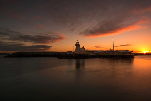 View of Howth Harbour lighthouse at sunset with a stone breakwater and calm water under a vibrant orange sky in Ireland. View of Howth Harbour lighthouse at sunset with a stone breakwater and calm water under a vibrant orange sky in Ireland.