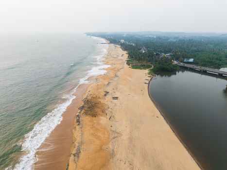 Aerial view of the sandy coastline where the ocean meets a calm river mouth surrounded by lush tropical palm trees under a hazy sky Galle Southern Province Sri Lanka.