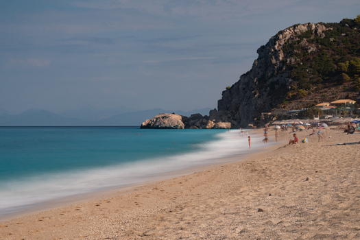 View of the azure sea kissing the sandy shore under a bright sky shadowed by a rocky cliff where people relax under umbrellas Lefkada Greece. View of the azure sea kissing the sandy shore under a bright sky shadowed by a rocky cliff where people relax under umbrellas Lefkada Greece.