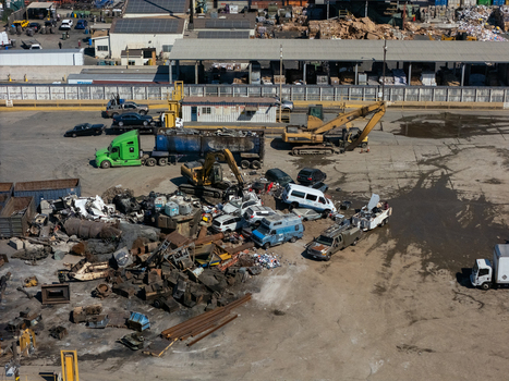 Aerial view of a scrap metal recycling yard featuring excavators piles of crushed cars and industrial trucks in Carson California United States.