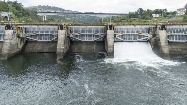 Aerial view of the Velle Dam with water flowing through a spillway gate on the Mino River surrounded by green hills and a bridge in Ourense Ourense Spain.