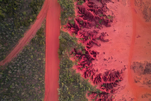 Aerial view of a vibrant red dirt road cutting through the rugged outback landscape with deep crimson gullies and sparse green vegetation in Broome Western Australia Australia.