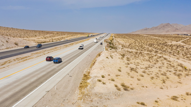 Aerial view of a highway stretching through a vast desert landscape with sparse vegetation and distant mountains under a clear blue sky in the United States.