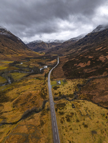 Aerial view of a long asphalt road winding through a dramatic mountain valley with snow capped peaks under a cloudy sky Scotland United Kingdom. Aerial view of a long asphalt road winding through a dramatic mountain valley with snow capped peaks under a cloudy sky Scotland United Kingdom.
