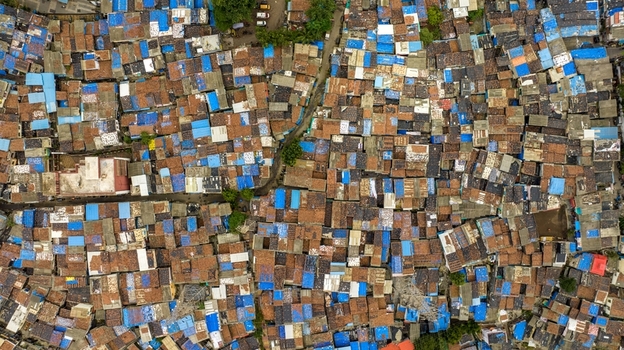 Aerial view of the dense urban fabric of Dharavi Slums featuring a complex network of rooftops with blue tarpaulins and brown tiles and narrow alleyways in Mumbai Maharashtra India. Aerial view of the dense urban fabric of Dharavi Slums featuring a complex network of rooftops with blue tarpaulins and brown tiles and narrow alleyways in Mumbai Maharashtra India.