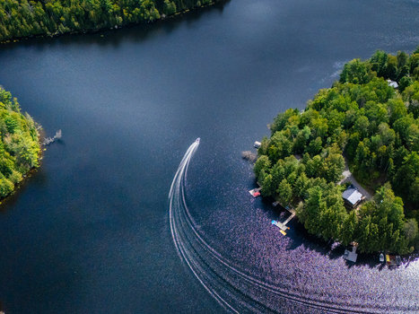 Aerial view of a motorboat leaving a white wake on the dark blue water of Baptiste Lake surrounded by forests and small docks along the shoreline in Hastings Highlands Ontario Canada.