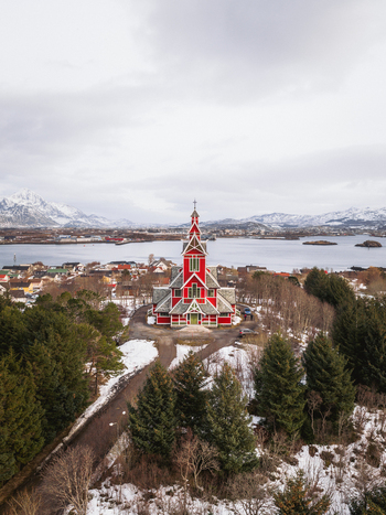 Aerial view of a striking red church standing proudly amidst the green pines and lingering snow with the serene waters and distant mountains painting a vivid backdrop Gravdal Nordland Norway. Aerial view of a striking red church standing proudly amidst the green pines and lingering snow with the serene waters and distant mountains painting a vivid backdrop Gravdal Nordland Norway.