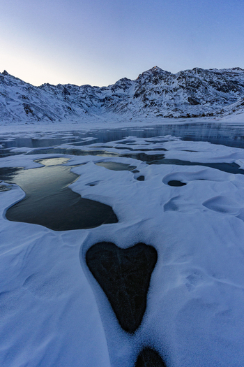 View of a heart shaped ice patch on a frozen lake surrounded by snow covered mountains at dusk in Madesimo Lombardy Italy. View of a heart shaped ice patch on a frozen lake surrounded by snow covered mountains at dusk in Madesimo Lombardy Italy.