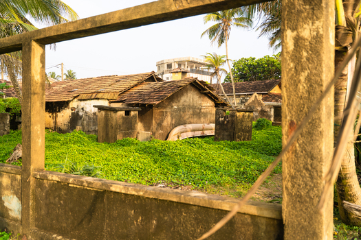 View of weathered traditional houses with tiled roofs surrounded by lush green vegetation and palm trees seen through a concrete frame in Galle Southern Province Sri Lanka.