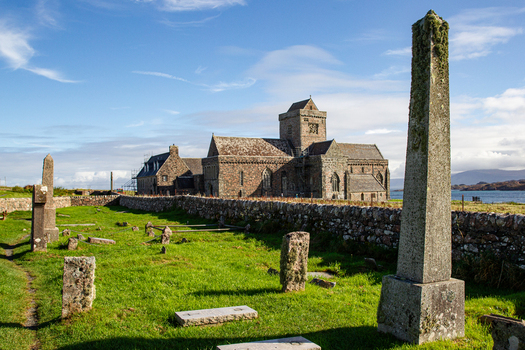 View of Iona Abbey and Nunnery featuring historic stone architecture an ancient graveyard with obelisks and a low stone wall under a bright blue sky Isle of Iona Scotland United Kingdom. View of Iona Abbey and Nunnery featuring historic stone architecture an ancient graveyard with obelisks and a low stone wall under a bright blue sky Isle of Iona Scotland United Kingdom.