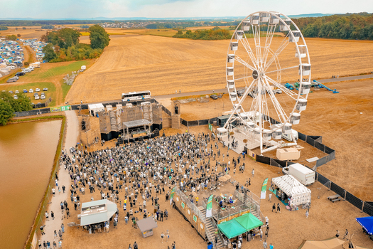 Herzogenaurach Germany 22 July 2022 Aerial view of the Open Beatz Festival featuring a large white Ferris wheel a stage and a crowd of people surrounded by golden fields. Herzogenaurach Germany 22 July 2022 Aerial view of the Open Beatz Festival featuring a large white Ferris wheel a stage and a crowd of people surrounded by golden fields.