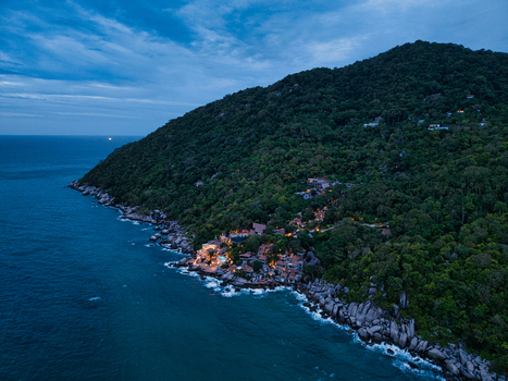 Aerial view of the rocky coastline of Ko Tao at dusk with illuminated resort buildings nestled in dense forest and rugged cliffs overlooking the sea Ko Tao Surat Thani Thailand.