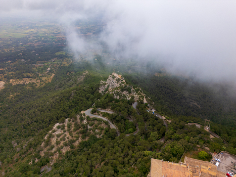 Aerial view of the Sanctuary of Sant Salvador on a mountain peak with winding roads through dense forest under thick white clouds in Felanitx Illes Balears Spain. Aerial view of the Sanctuary of Sant Salvador on a mountain peak with winding roads through dense forest under thick white clouds in Felanitx Illes Balears Spain.