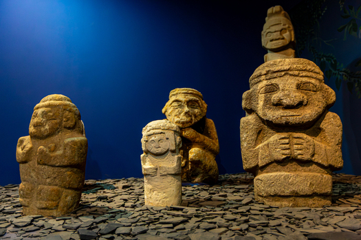 View of ancient stone statues at San Agustin Archaeological Park displayed on a bed of dark stone fragments against a blue background in San Agustin Huila Colombia.