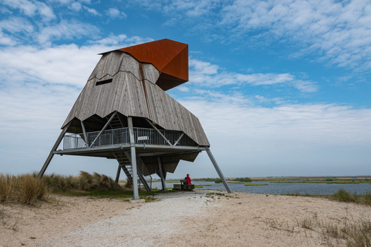 View of the Marker Wadden bird watching tower with a person sitting on a bench nearby set against a blue sky and water in Lelystad Flevoland Netherlands. View of the Marker Wadden bird watching tower with a person sitting on a bench nearby set against a blue sky and water in Lelystad Flevoland Netherlands.
