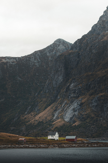 View of a small white house and a red shed nestled at the base of a towering rugged mountain by the water in Ramberg Nordland Norway. View of a small white house and a red shed nestled at the base of a towering rugged mountain by the water in Ramberg Nordland Norway.