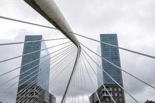 View of the white cables and arch of the Zubizuri bridge with the modern Isozaki Atea glass towers in the background under a cloudy sky in Greater Bilbao Basque Country Spain. View of the white cables and arch of the Zubizuri bridge with the modern Isozaki Atea glass towers in the background under a cloudy sky in Greater Bilbao Basque Country Spain.