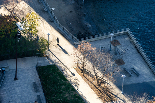 Manhattan United States 01 December 2025 View of a lone figure strolling along the East River waterfront bisected by sharp sunlight and shadows where the city meets the tranquil water s edge.