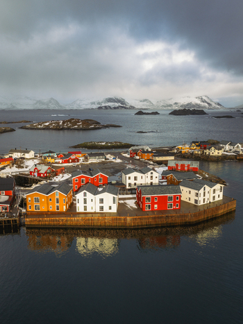 Aerial view of vibrant red white and yellow cabins clustered on a small island in a dark harbor with snow dusted mountains in the distance Henningsv&aelig;r Nordland Norway.