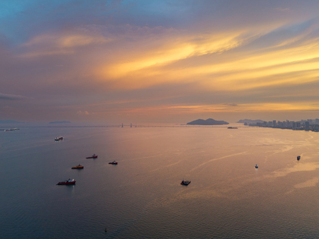 Aerial view of the Penang Bridge spanning across the sea with several ships anchored nearby under a vibrant sunset sky in George Town Penang Malaysia.