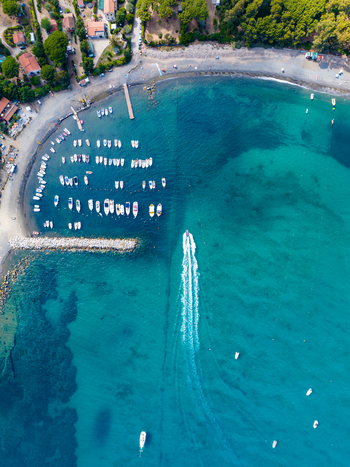 Aerial view of the turquoise waters of Lacona bay with a small boat leaving a wake moored vessels near the pier and the sandy coastline in Lacona Tuscany Italy.