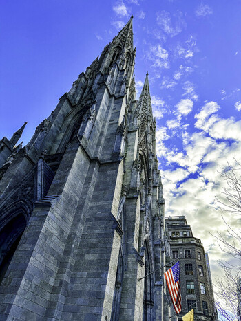 View of St. Patrick s Cathedral on Fifth Avenue Midtown Manhattan featuring Gothic revival stonework spires and an American flag against a blue sky in New York New York United States.