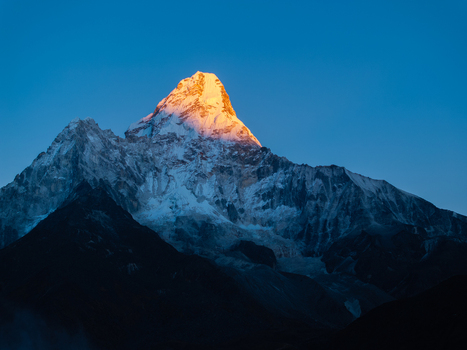 Aerial view of the snow capped Himalayan mountain peak illuminated by golden hour sunlight against a clear deep blue sky in Nepal.