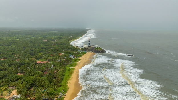 Aerial view of Kapu Light House standing on a rocky outcrop along the sandy coastline with lush palm trees and crashing waves in Udupi Karnataka India.