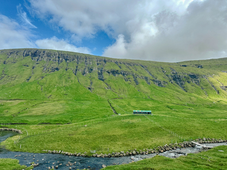 View of a small green building nestled in the lush grassy hills of the Faroe Islands with a stream flowing through the foreground under a cloudy sky Faroe Islands. View of a small green building nestled in the lush grassy hills of the Faroe Islands with a stream flowing through the foreground under a cloudy sky Faroe Islands.