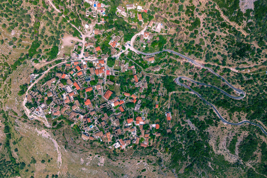 Aerial view of the historic stone houses and red rooftops of a hillside village with a winding road through lush greenery in Qeparo Fshat Vlore County Albania. Aerial view of the historic stone houses and red rooftops of a hillside village with a winding road through lush greenery in Qeparo Fshat Vlore County Albania.