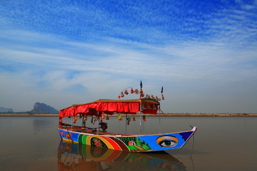 View of a colorful decorated boat with a red canopy floating on the calm waters of the Chenab River under a bright blue sky in Chiniot Punjab Pakistan.