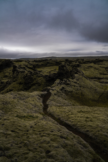 View of a mossy rugged landscape under a brooding sky where a narrow trail carves through the textured terrain Skaftárhreppur Iceland. View of a mossy rugged landscape under a brooding sky where a narrow trail carves through the textured terrain Skaftárhreppur Iceland.