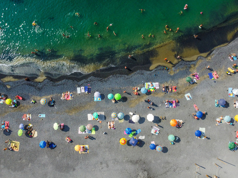 Aerial view of people sunbathing under colorful umbrellas on a pebble beach and swimming in turquoise water in San Lorenzo Liguria Italy.