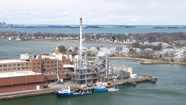 Aerial view of an industrial complex with brick buildings metallic structures and a tall chimney casting a shadow onto the pier where boats are docked Quincy Massachusetts United States.