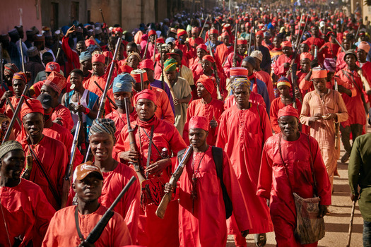 Zaria Nigeria 21 March 2026 View of a vibrant procession of people adorned in striking red garments carrying traditional rifles moving through sun drenched streets. Zaria Nigeria 21 March 2026 View of a vibrant procession of people adorned in striking red garments carrying traditional rifles moving through sun drenched streets.