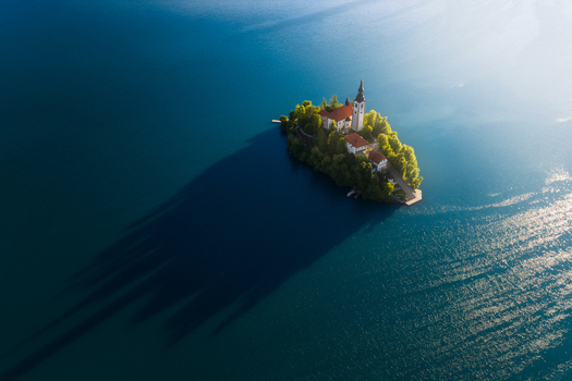 Aerial view of Lake Bled island featuring the Pilgrimage Church of Mary surrounded by lush trees and a long shadow on the blue water Bled Radovljica Slovenia.