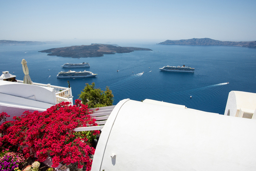 View of Fira with vibrant red bougainvillea flowers in the foreground overlooking the blue Aegean Sea and cruise ships in the caldera in Fira Greece. View of Fira with vibrant red bougainvillea flowers in the foreground overlooking the blue Aegean Sea and cruise ships in the caldera in Fira Greece.