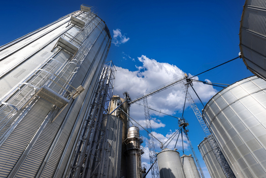 View of tall silver industrial grain silos and metal storage elevators under a bright blue sky with white clouds in Toronto Ontario Canada.