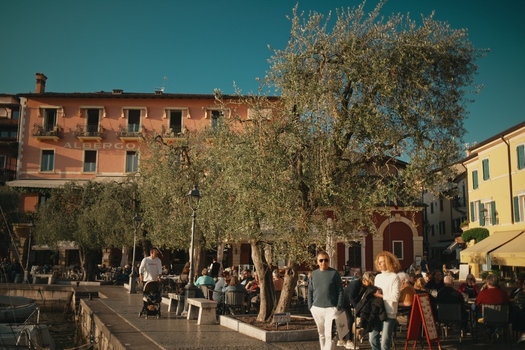 View of Lake Garda waterfront square with a large olive tree and people sitting at outdoor cafes Italy. View of Lake Garda waterfront square with a large olive tree and people sitting at outdoor cafes Italy.