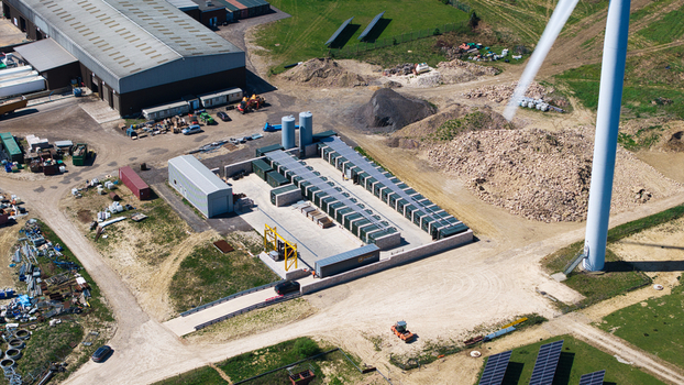 Aerial view of Chelveston Wind Farm featuring battery storage containers solar panels and a large wind turbine near industrial buildings in Wellingborough England United Kingdom. Aerial view of Chelveston Wind Farm featuring battery storage containers solar panels and a large wind turbine near industrial buildings in Wellingborough England United Kingdom.