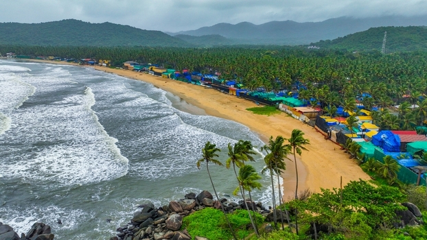 Aerial view of Palolem Beach with colorful huts nestled among palm trees along the sandy coastline crashing waves and distant mountains in Canacona Goa India.