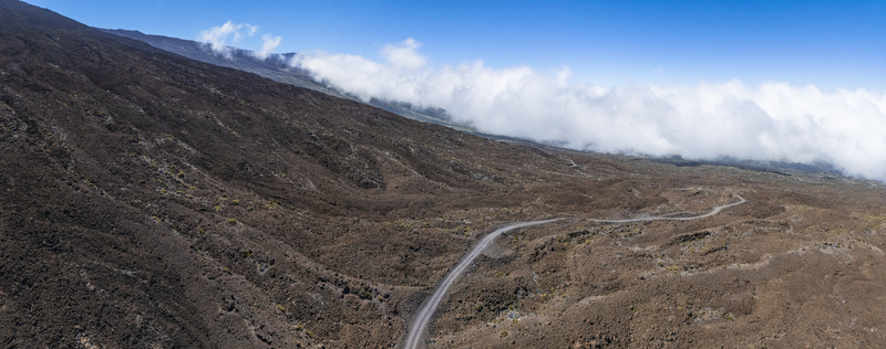 Aerial view of the rugged slopes of Mauna Kea featuring a winding dirt road through dark volcanic terrain and white clouds Hawaii County Hawaii United States.