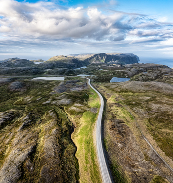 Aerial view of a winding road cutting through the rugged tundra landscape with rocky terrain small lakes and distant cliffs in Nordkapp Municipality Finnmark Norway. Aerial view of a winding road cutting through the rugged tundra landscape with rocky terrain small lakes and distant cliffs in Nordkapp Municipality Finnmark Norway.