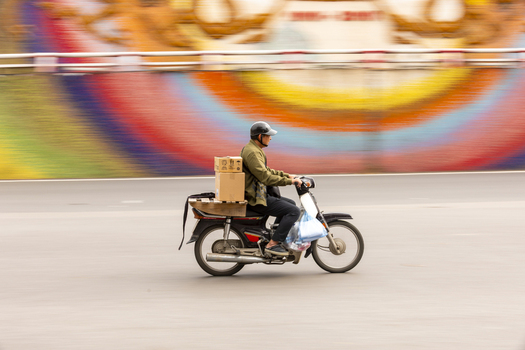 View of a delivery person riding a motorbike with cardboard boxes on the back against a colorful motion blurred background in Hanoi Ha Noi Vietnam.