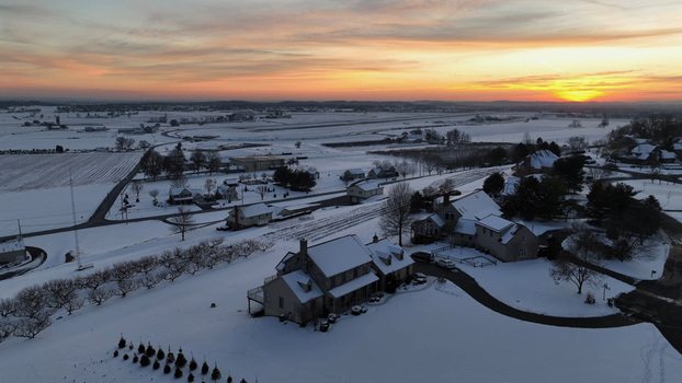 Aerial view of a snow covered landscape featuring residential houses bare trees and open fields under a vibrant sunset sky in Warwick Township Pennsylvania United States.
