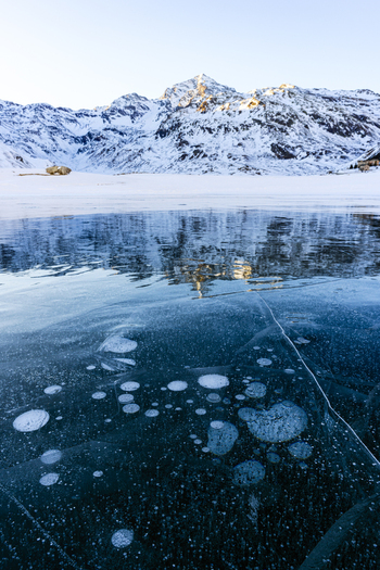 View of frozen lake with trapped air bubbles reflecting snow covered mountain peaks under a clear winter sky in Madesimo Lombardy Italy. View of frozen lake with trapped air bubbles reflecting snow covered mountain peaks under a clear winter sky in Madesimo Lombardy Italy.